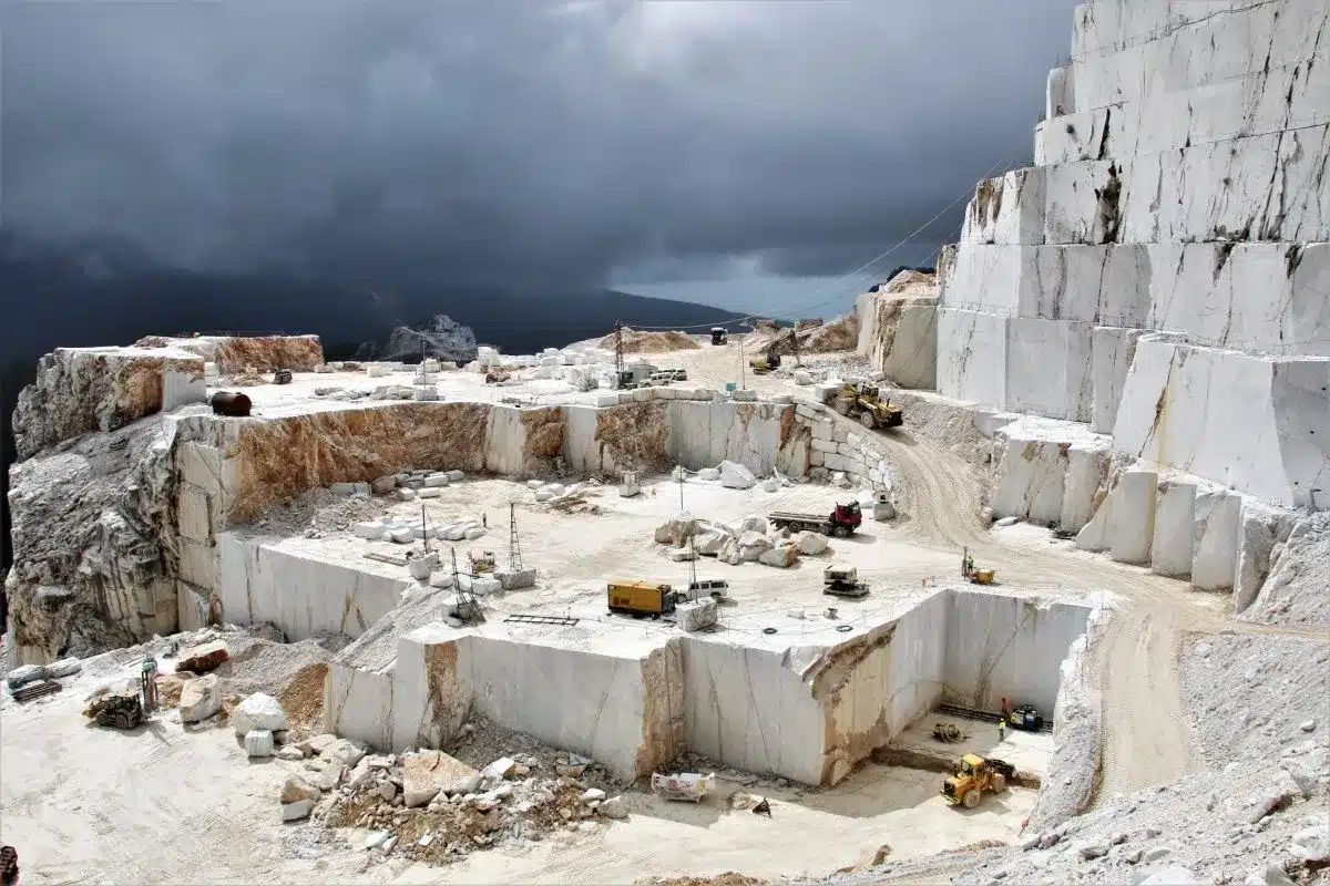 Carrara marble quarry with large white stone blocks, heavy machinery, and workers under a cloudy sky.