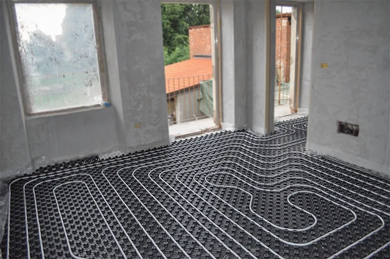 Underfloor heating pipes installed on black insulation panels in an unfinished room with large windows and door openings.