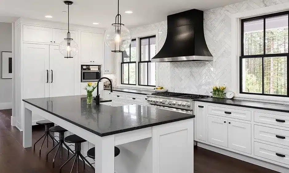 Modern kitchen with black marble countertops, white cabinetry, large island, and black range hood under large windows.