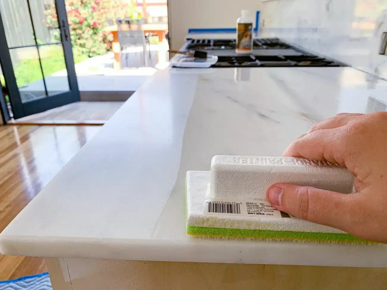 Hand applying sealant on a marble countertop with a sponge applicator in a bright kitchen setting.